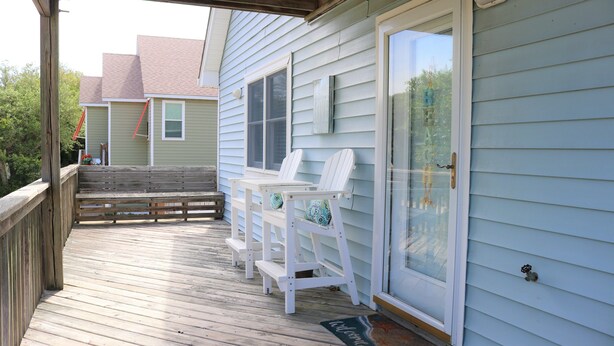 A wooden deck with white chairs and a bench, a sliding glass door, and a welcome mat. - Bright & Beachy 3 bed/2 bath Soundside MP6 (Kill Devil Hills)