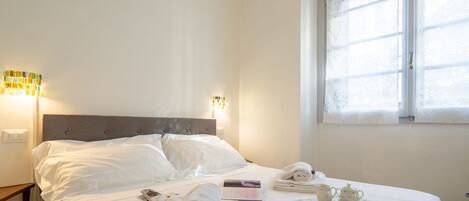 A neatly made bed with white linens, a bedside table with a lamp, and a window with floral curtains.