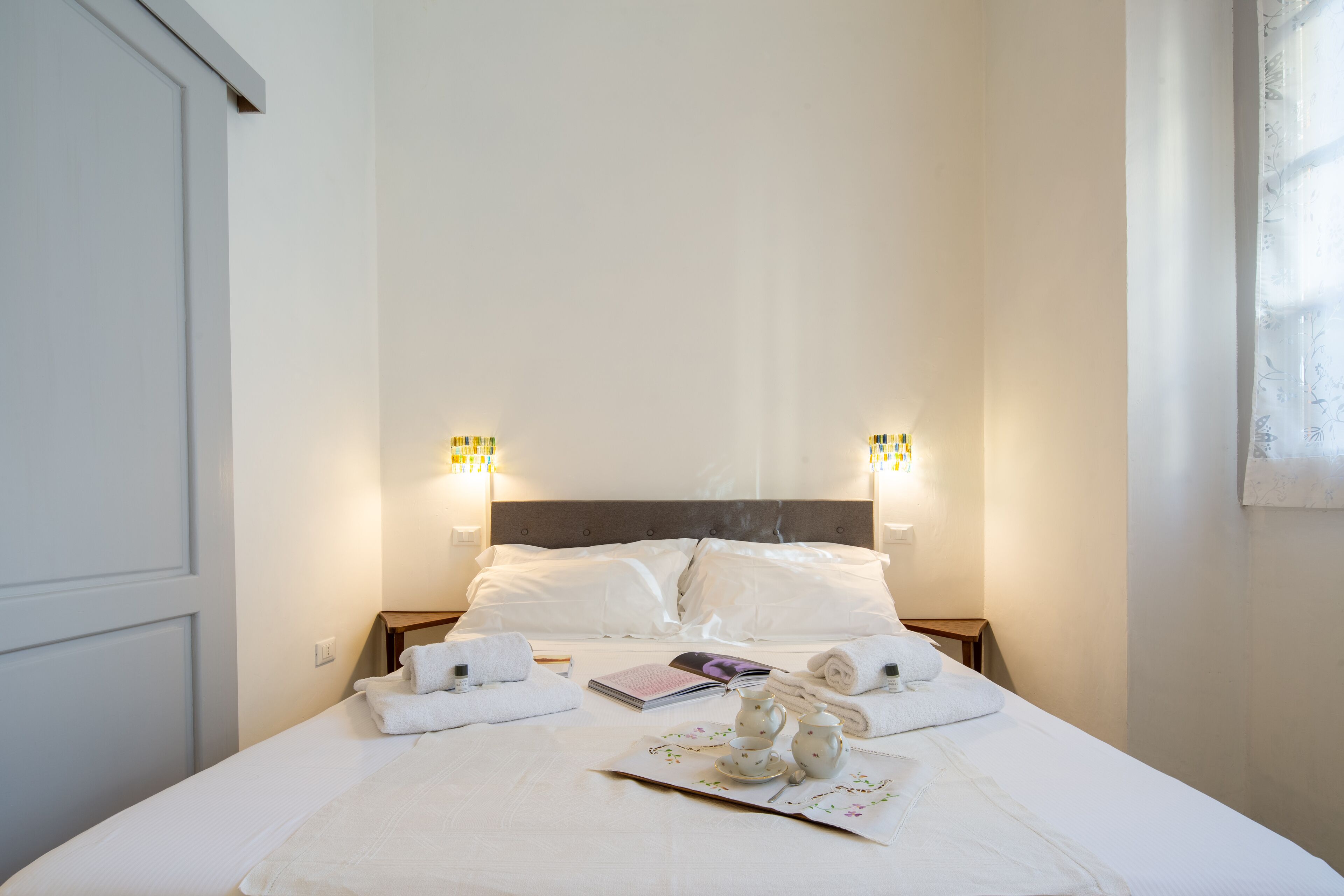 A neatly made bed with white linens, a bedside table with a book and a teapot, and two wall-mounted lamps.
