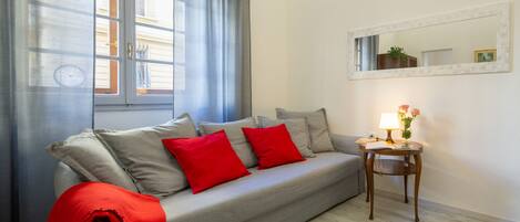A living room with a grey sofa, red cushions, a small wooden side table with a lamp, and a mirror on the wall.