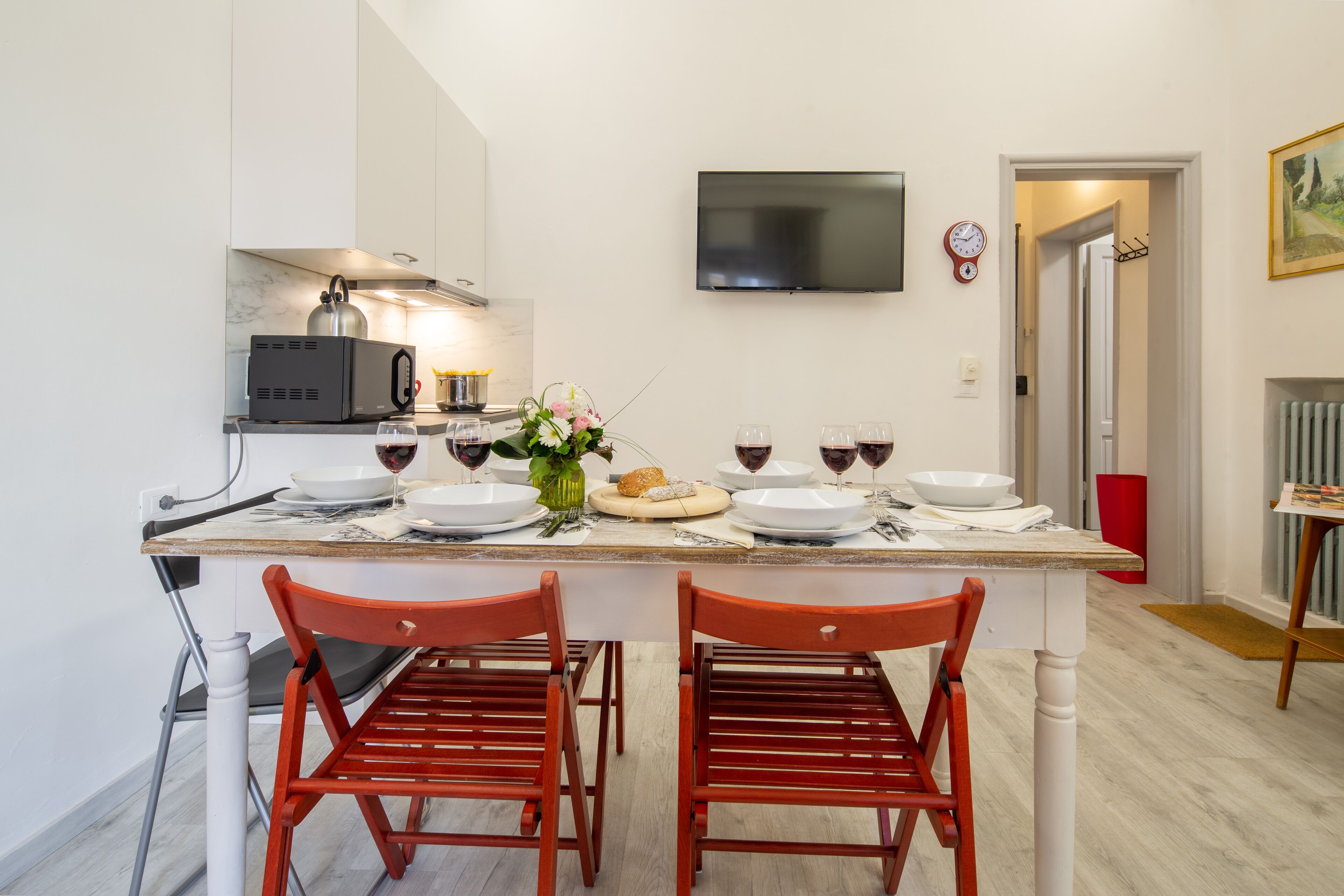 A modern kitchen with a dining table set for two, equipped with a microwave, kettle, and wine glasses.