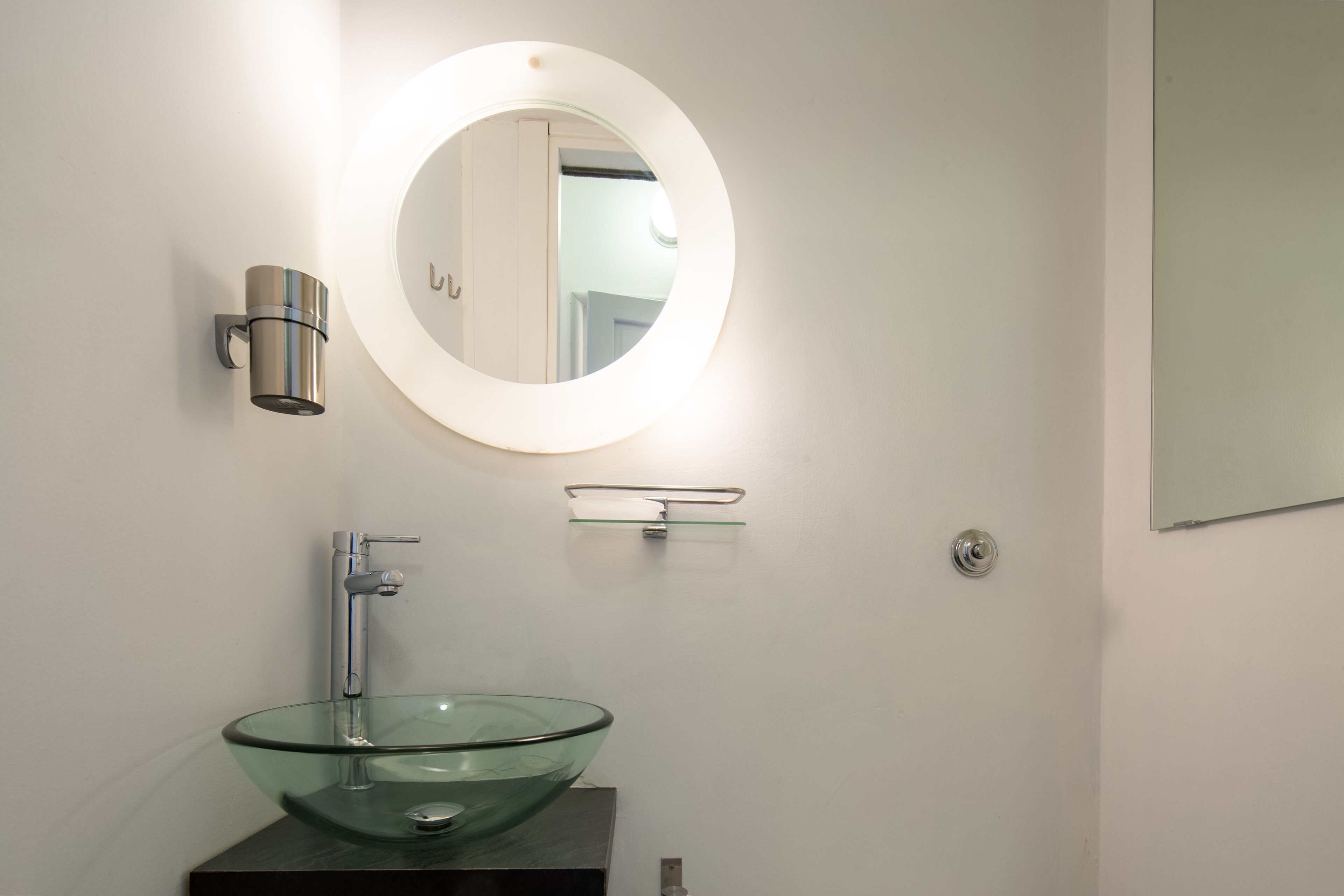 A bathroom with a glass bowl sink, a round mirror with a light, and a wall-mounted towel rack.