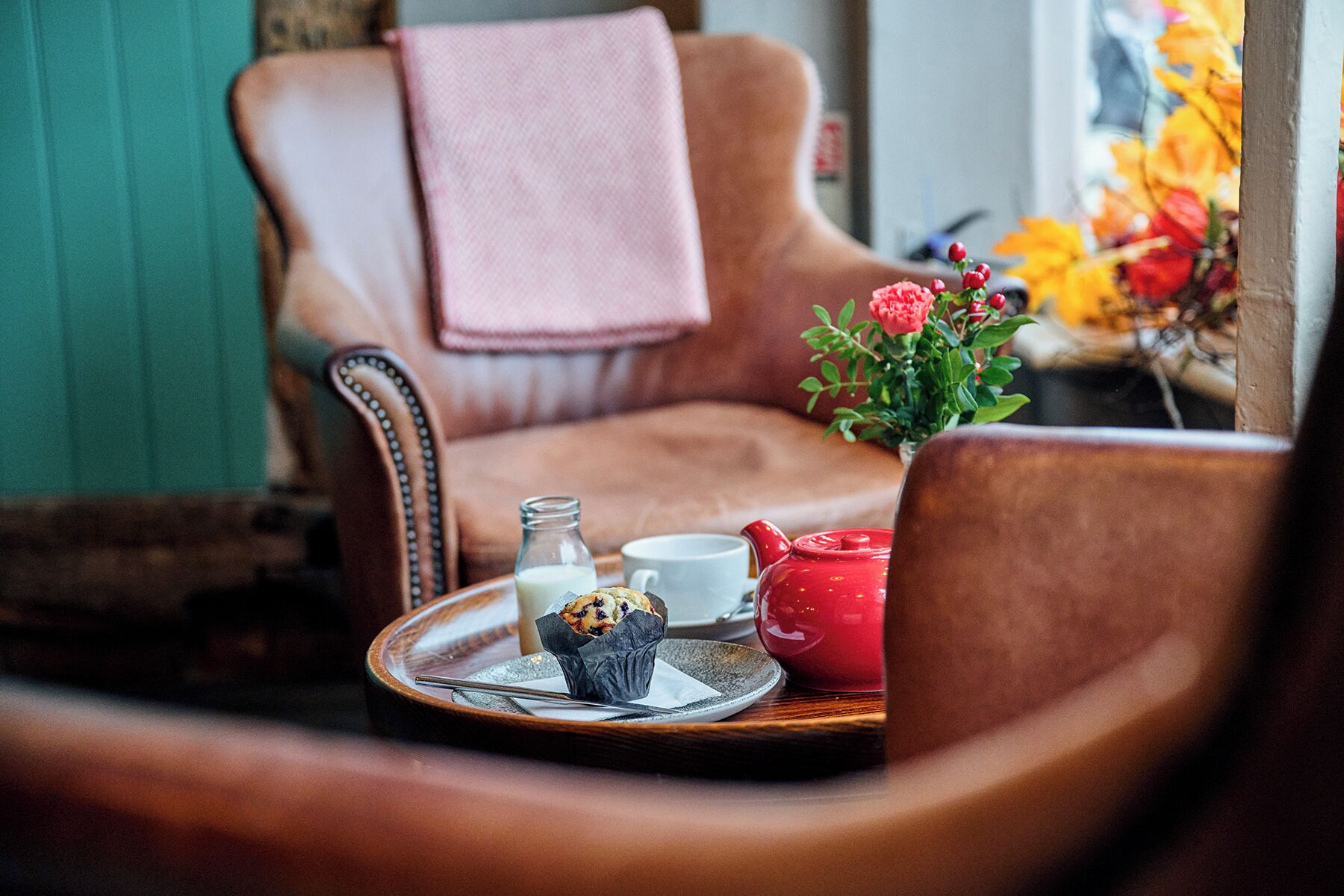 Un rincón acogedor con un sillón de cuero, una mesita con una bandeja que contiene una tetera, una taza y un muffin.