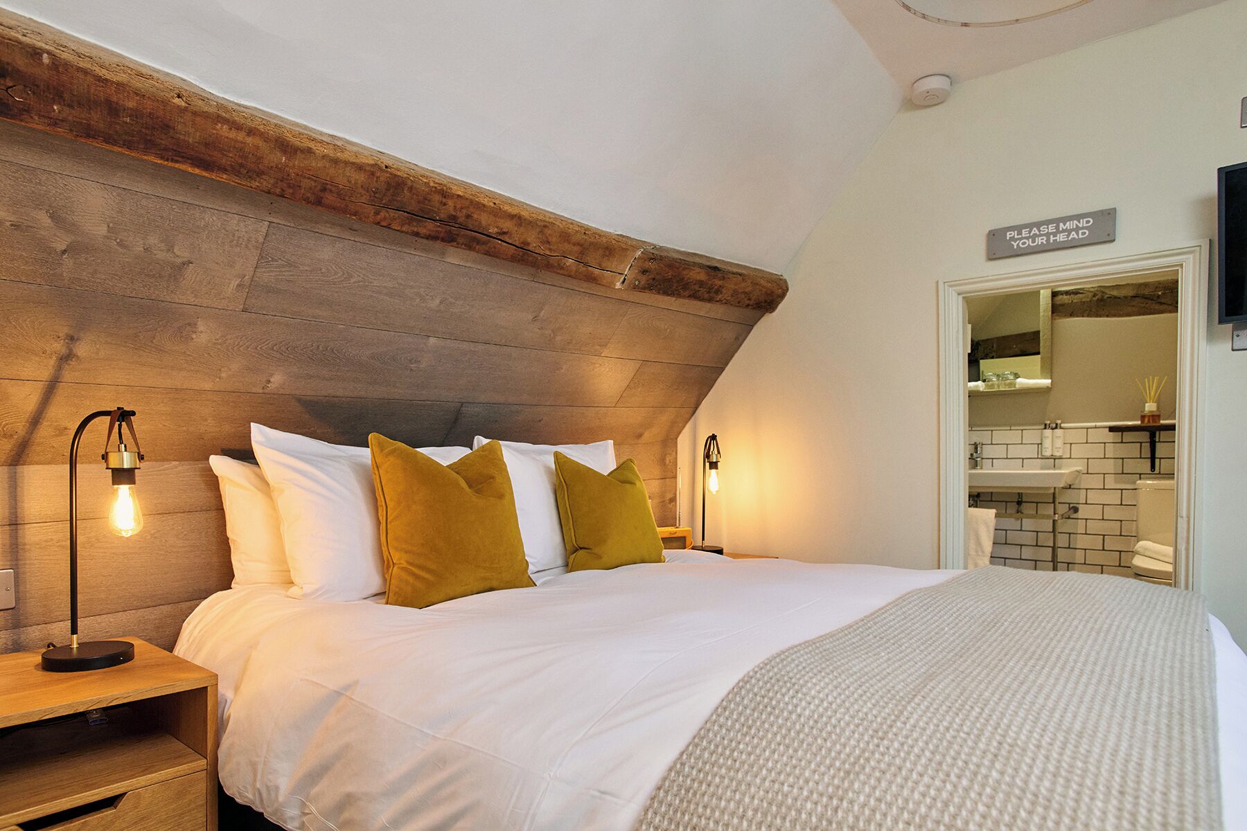 A bedroom with a wooden headboard, a bed with white bedding and mustard pillows, bedside lamps, and a bathroom visible through an open door.