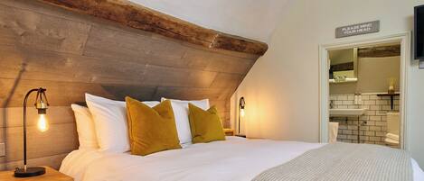 A bedroom with a wooden headboard, a bed with white bedding and mustard pillows, bedside lamps, and a bathroom visible through an open door.