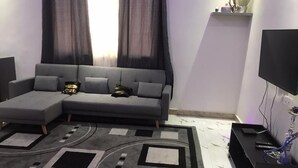 A modern living room with a grey sectional sofa, a patterned rug, a flat-screen TV, and a small shelf with decorative items. - F2 UDDERS AT MAYAA (Ngor)