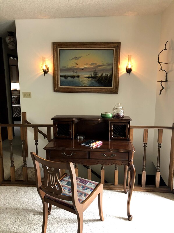 A wooden desk with a framed painting above it, two wall sconces, and a chair with a patterned cushion.