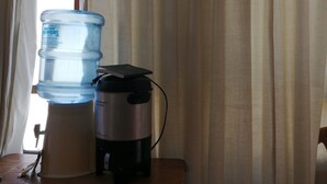 A water dispenser and a coffee maker on a wooden table near a window with curtains. - Posada el mesón xela (Quetzaltenango)