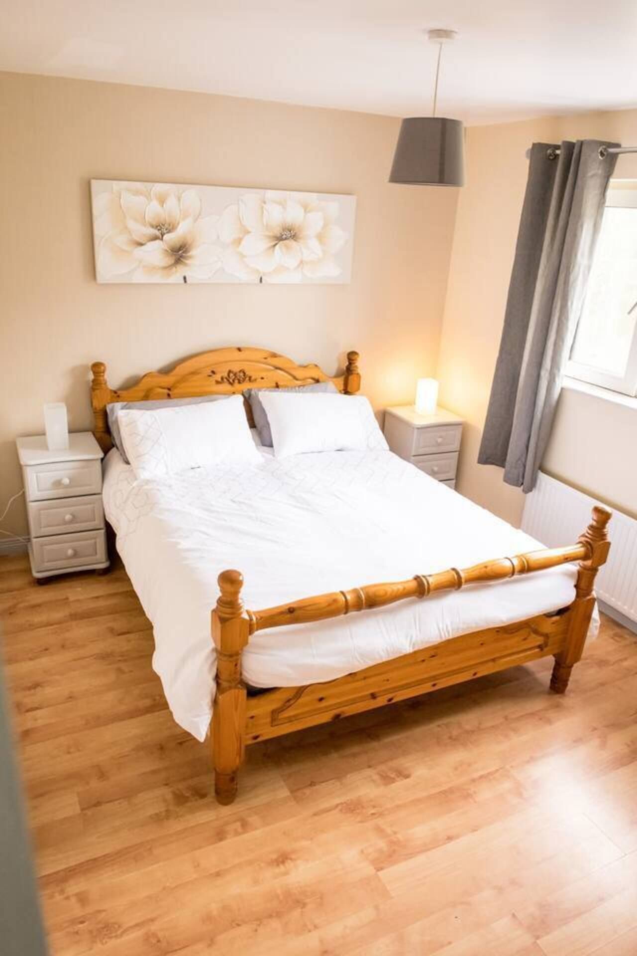 Una cama de madera con ropa de cama blanca en una habitación con suelo de madera, una mesita de noche, una lámpara de pared y una ventana con cortinas.