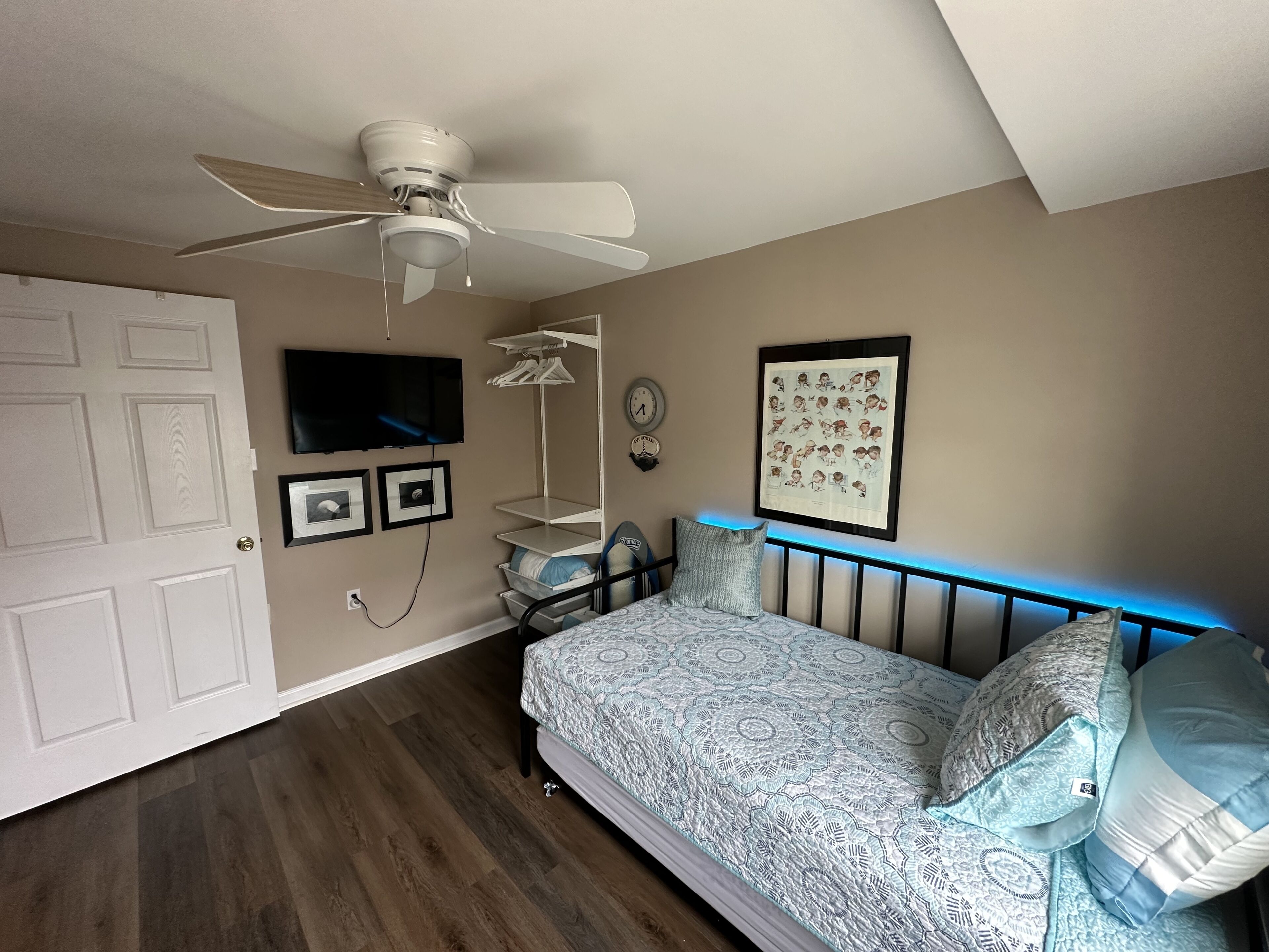Un dormitorio con cama, ventilador de techo, televisor, reloj de pared y un cuadro enmarcado.