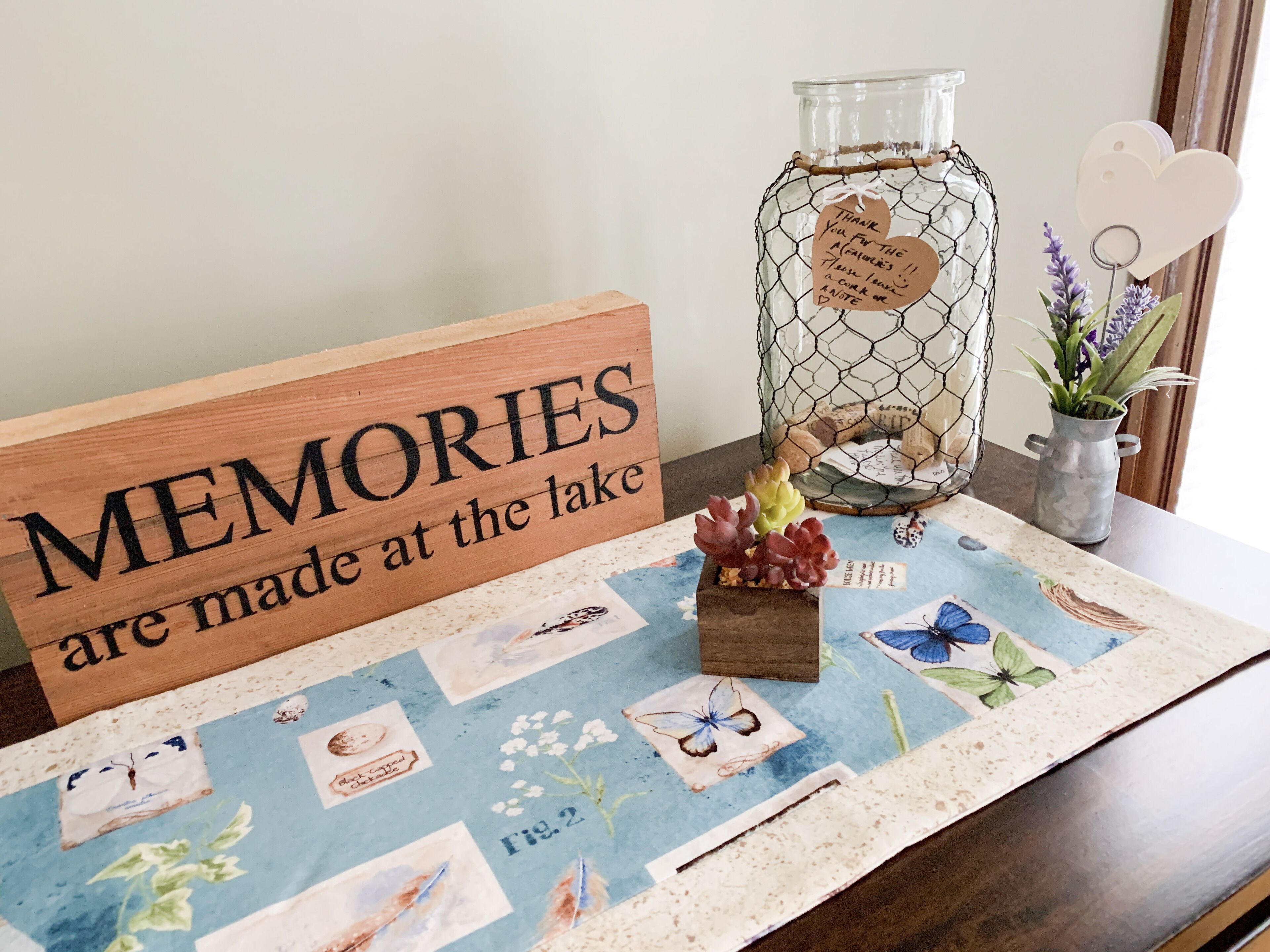 A wooden sign with the text "MEMORIES are made at the lake" and a decorative tablecloth with various illustrations.