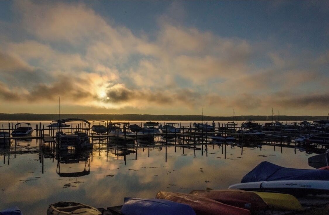 A marina with boats docked at wooden piers, calm water reflecting the sky, and a distant shoreline.