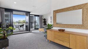 Une chambre d’hôtel moderne dotée d’une grande porte-fenêtre coulissante donnant sur un balcon, d’une console en bois, d’un grand miroir et d’un sol recouvert d’un tapis gris.