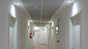 A long, narrow hallway with white walls and a speckled floor. There are doors on both sides, and a fire alarm is visible on the left wall.