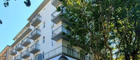 Un edificio de varios pisos con balcones, una tienda en la planta baja con diversos artículos a la vista, y árboles en primer plano.