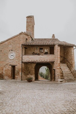 A rustic building with a brick facade, a clock, and a staircase leading to a balcony. - La suite in piazza in Borgo Loretello (Loretello)