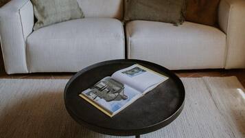 Un salon chaleureux avec un canapé de couleur claire, une table basse ronde noire sur laquelle repose un magazine, et un mur en briques.
