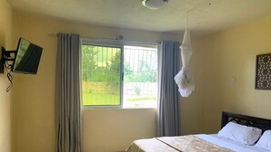 A hotel room with a bed, a TV mounted on the wall, a window with curtains, and a hanging light fixture. - Zaburi Retreat (Nguluni)