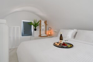 Un dormitorio ordenado con cama, una mesita de noche con una botella y vasos, una bandeja con fruta, una planta, y un estante en la pared con una toalla y una lámpara.