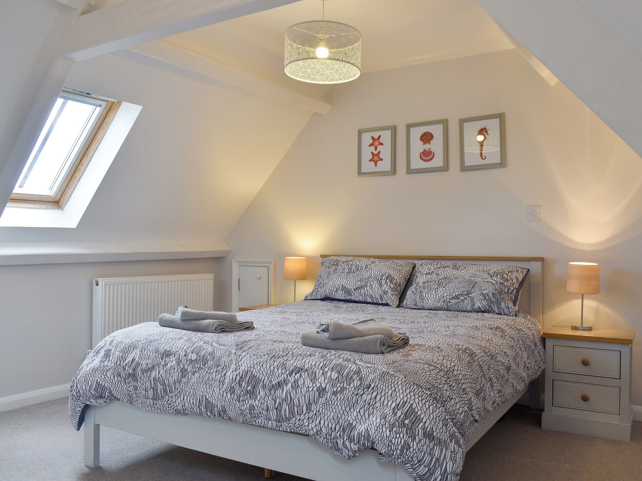 A bedroom with a sloped ceiling, a bed with patterned bedding, two bedside lamps, a radiator, and a skylight.