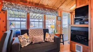 L’intérieur chaleureux d’un chalet avec un canapé, des coussins à motifs, une cheminée et de grandes fenêtres donnant sur un paysage pittoresque.