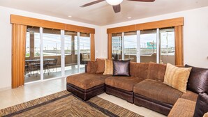 Amplia sala de estar con sofá modular, ventilador de techo y ventanas grandes con vistas al exterior.