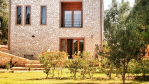 Una casa moderna de dos pisos de piedra, con puerta y ventanas de madera, rodeada por un jardín cuidado con árboles pequeños y un césped.