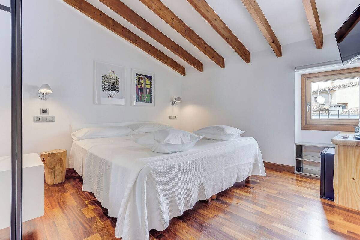Une chambre avec un plafond en bois, un lit recouvert de linge de lit blanc, deux lampes fixées au mur et une fenêtre donnant sur l’extérieur.