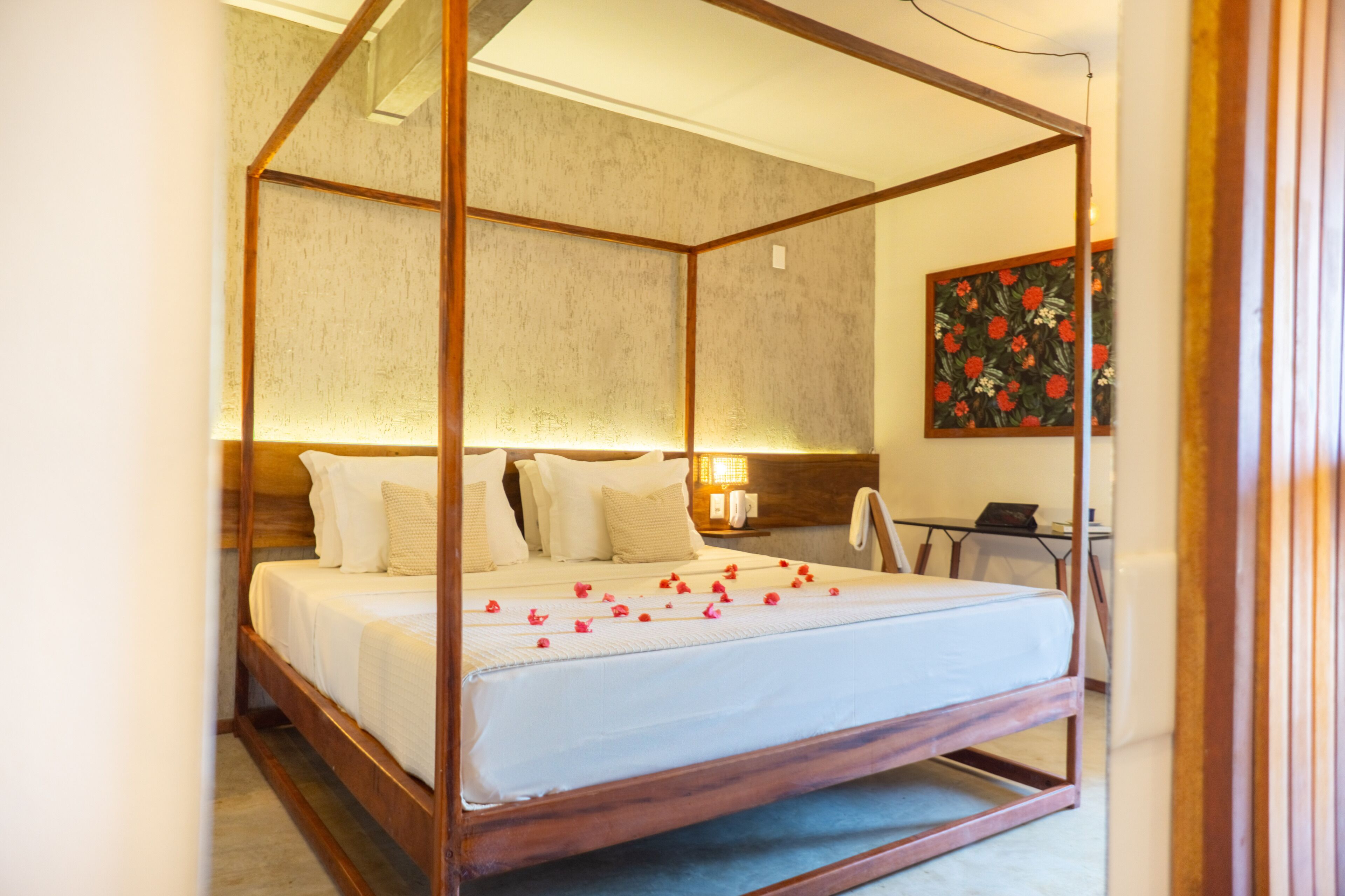 A four-poster bed with a wooden frame and white bedding, a bedside table with a lamp, and a floral painting on the wall.