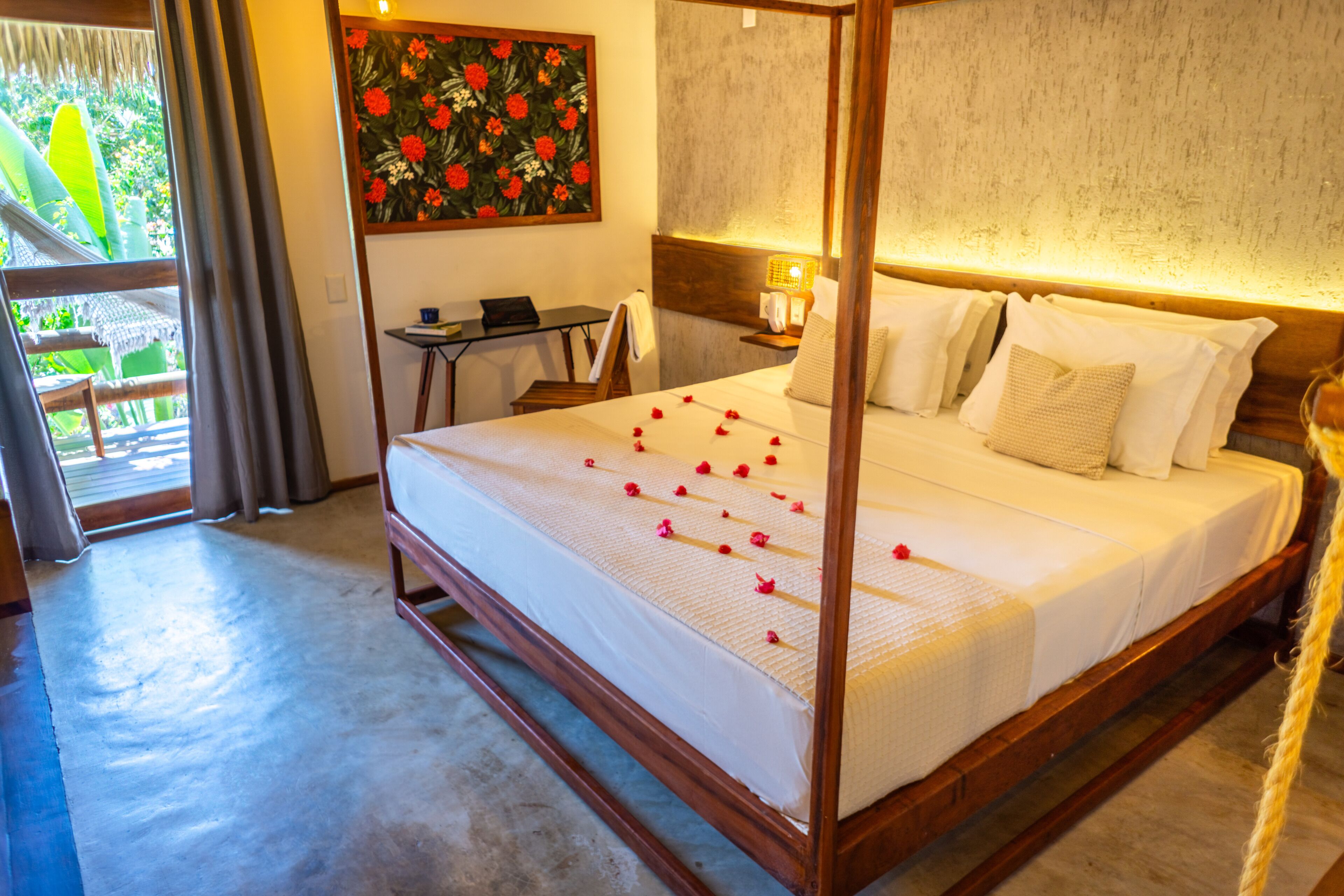 Een hemelbed met rozenblaadjes, een klein tafeltje met een laptop, en een bloemen schilderij aan de muur.