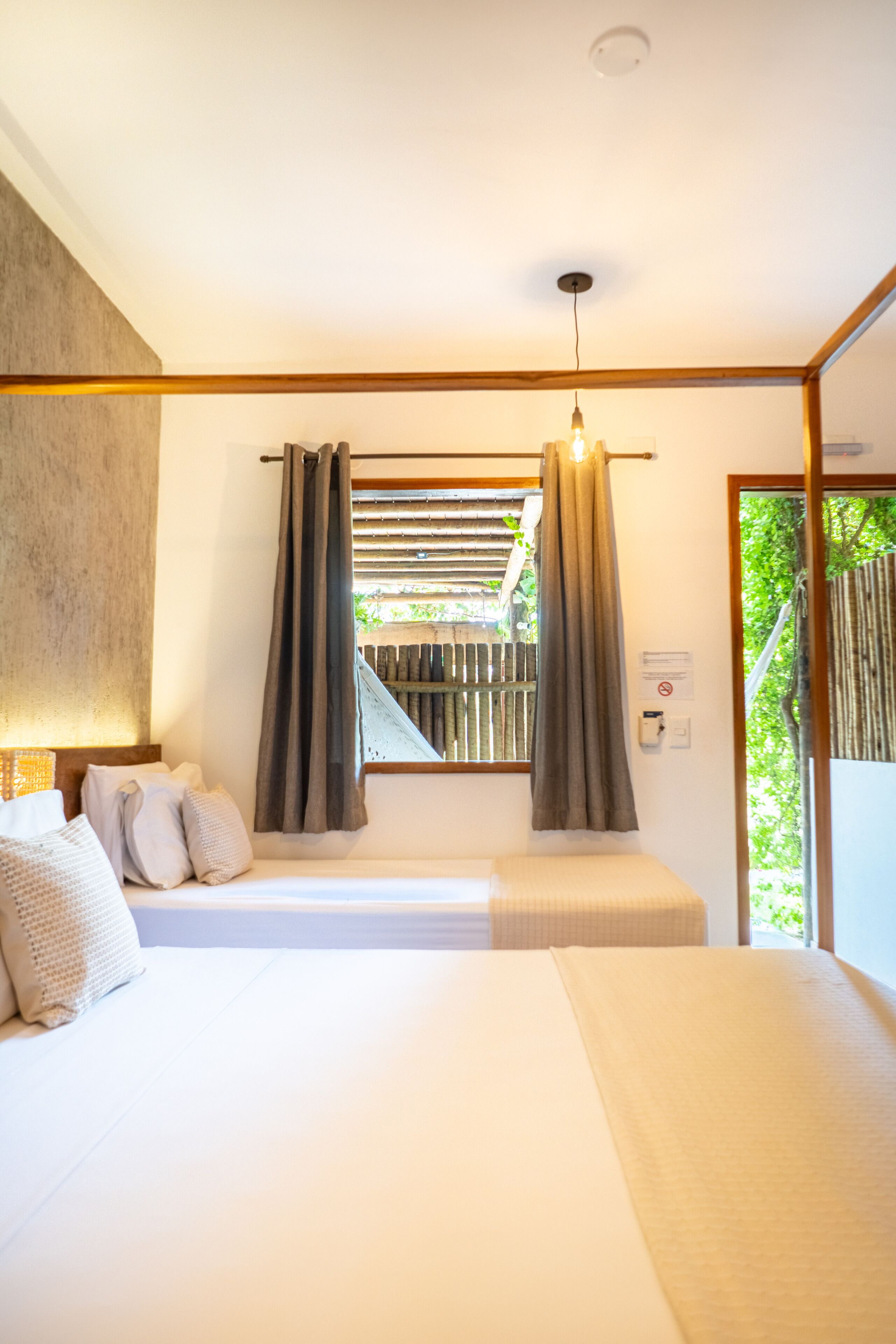 Een slaapkamer met een bed, kussens, een raam met gordijnen en uitzicht op groen.