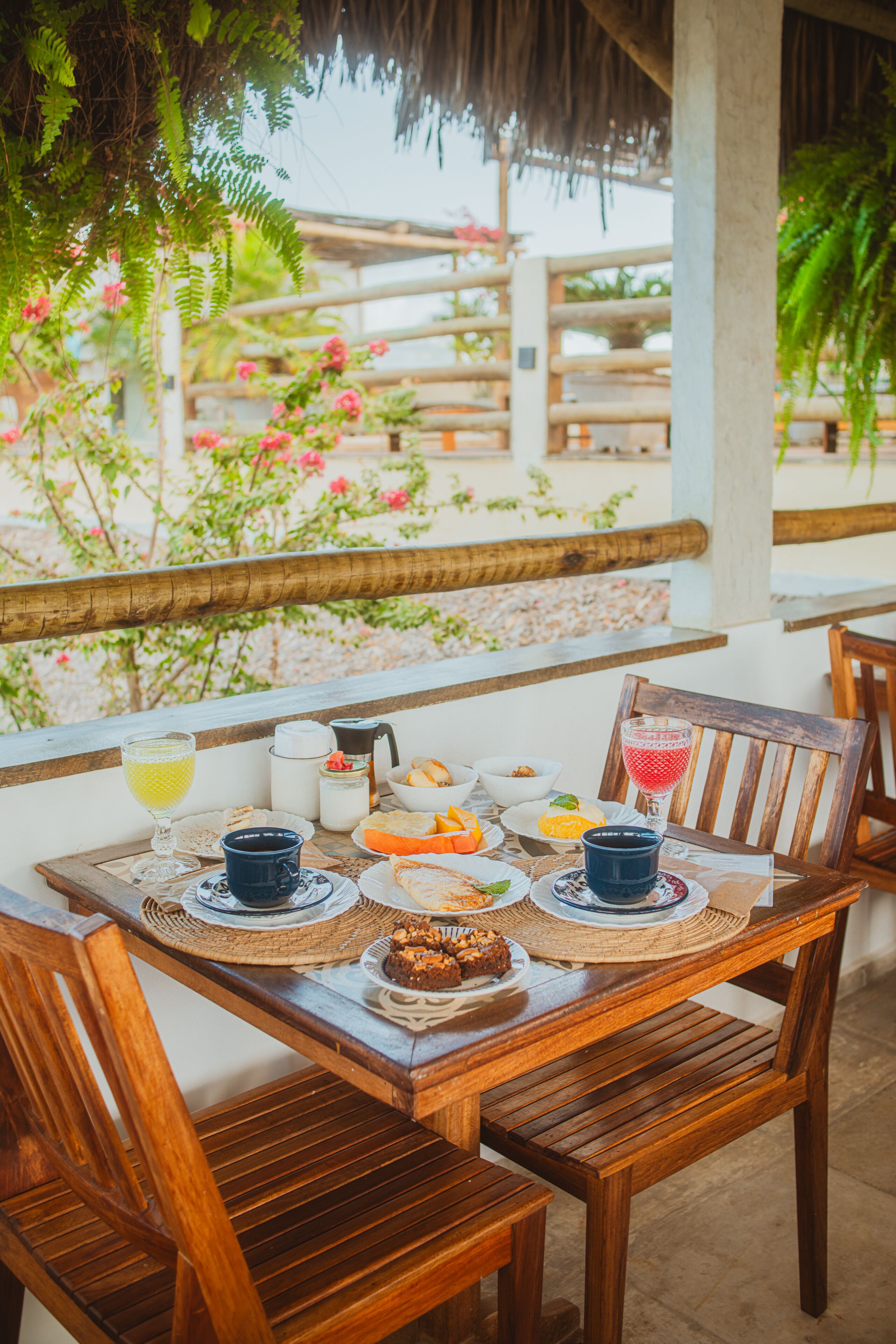 Una mesa de madera dispuesta con cosas para el desayuno, como café, zumo de naranja y pasteles, bajo un techo de paja con vista a un jardín.