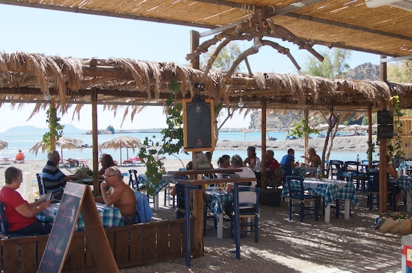 An outdoor dining area with thatched roof, tables, and chairs, overlooking a beach with umbrellas and people.