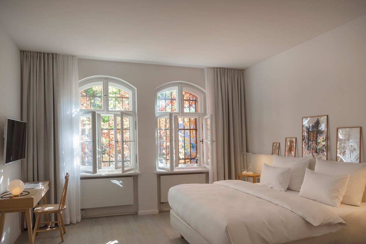 A bedroom with a bed, two windows with curtains, a desk with a chair, and a TV.