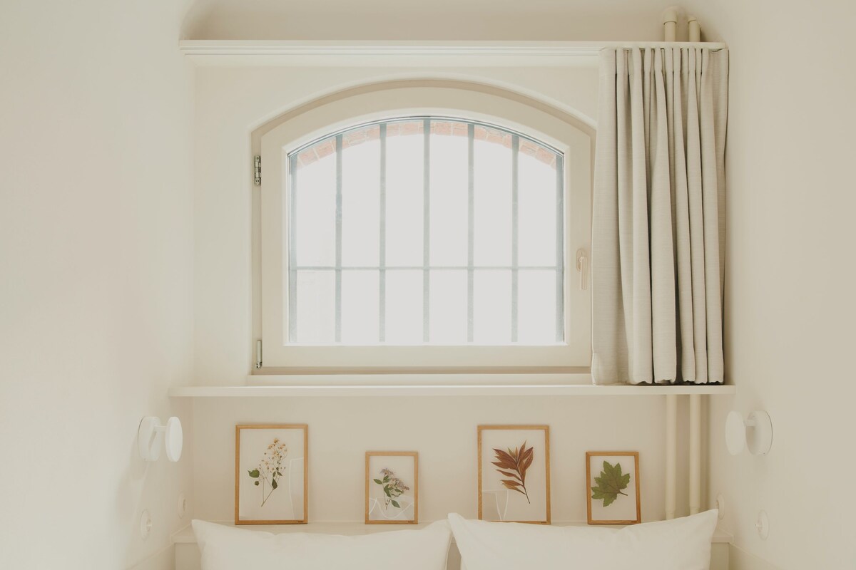 A bed with white bedding, a window with curtains, and three framed botanical prints on the wall.