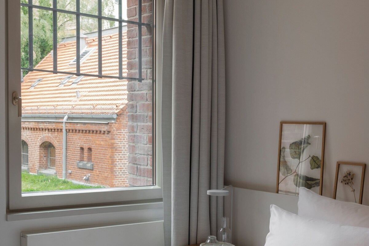 Une chambre avec une grande fenêtre, un lit recouvert de draps blancs, une petite table de chevet avec une lampe, et des tableaux encadrés accrochés au mur.