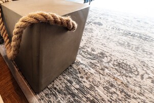 Una esquina de una habitación con una alfombra texturizada, un objeto grande y gris con forma de bloque, y una cuerda atada a él.