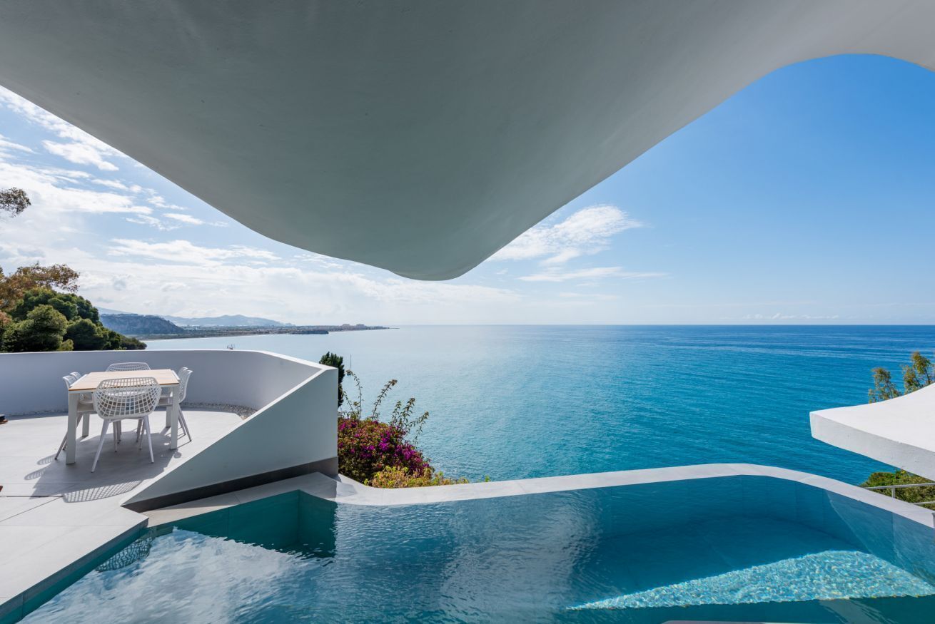 A modern outdoor pool with a view of the sea, a white table with chairs, and a curved white structure.