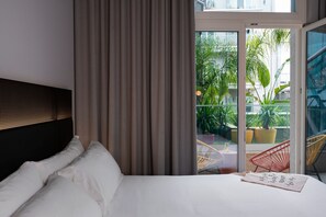 Quarto de hotel com cama, cortinas e vista para uma varanda com plantas e cadeiras.