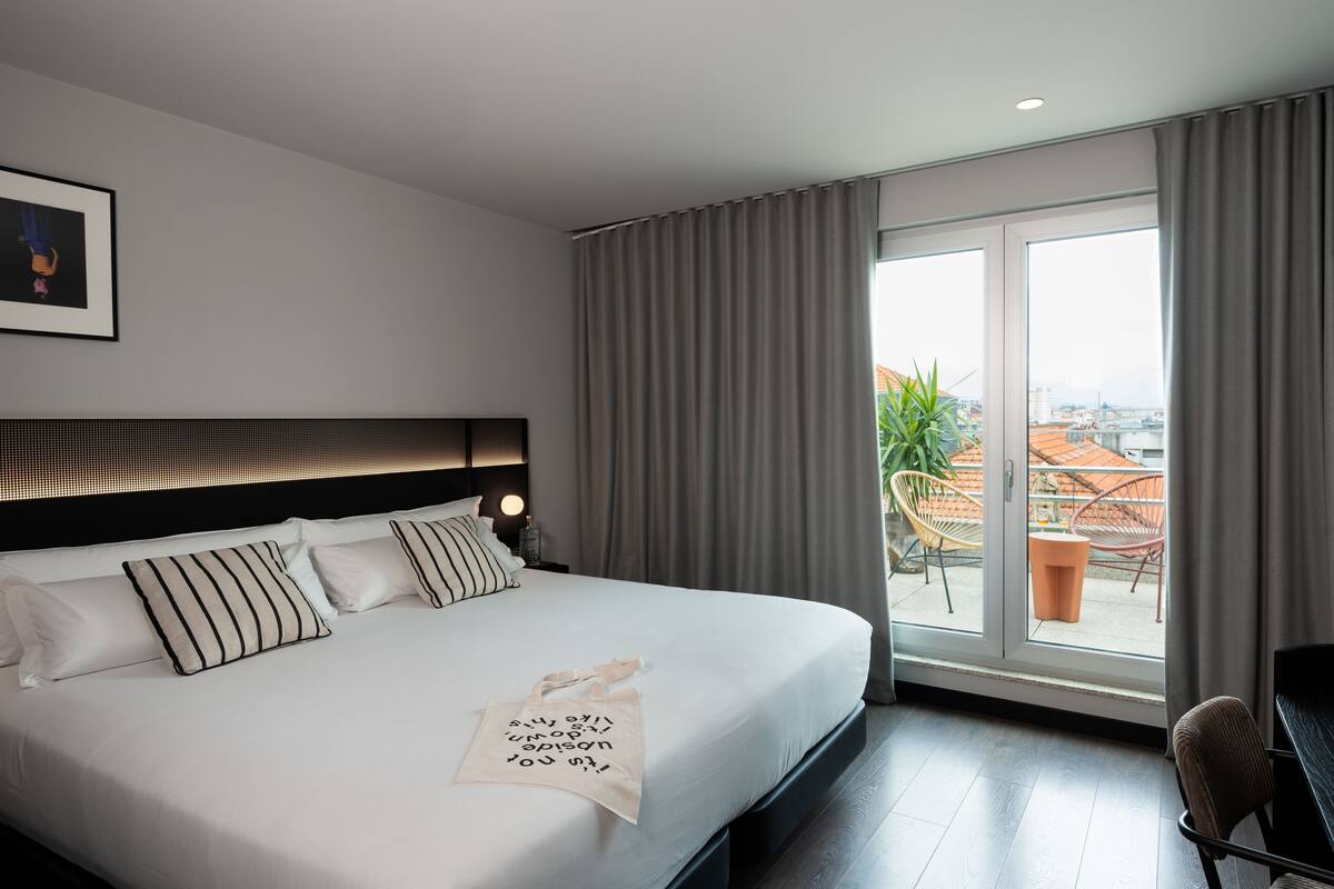 A modern hotel room with a large bed, a desk, and a balcony with a city view.