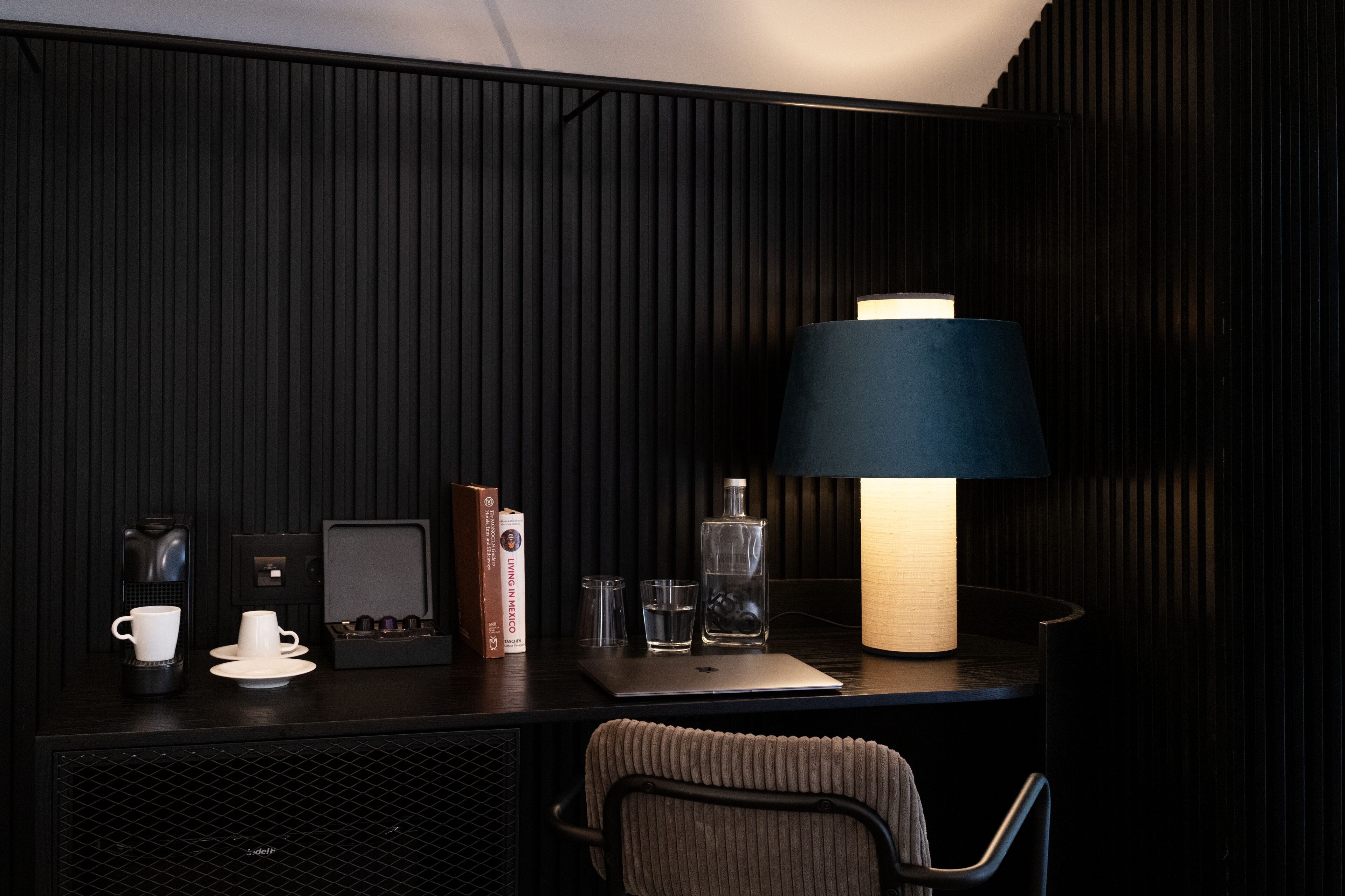 A modern hotel room with a dark wood panel wall, a desk with a laptop, a coffee maker, a book, a glass of water, and a lamp.
