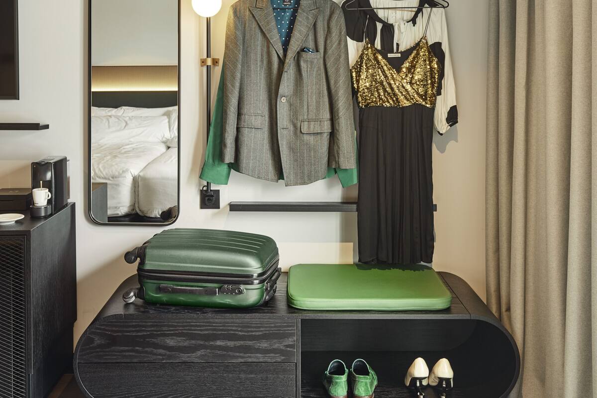 A hotel room with a bed, a dresser, a mirror, a coat rack with a suit and a dress, a suitcase, and a pair of shoes.