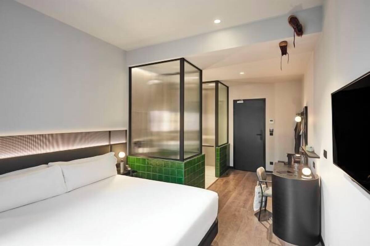 A modern hotel room with a bed, a glass partitioned shower area, a desk with a chair, and a mounted TV.