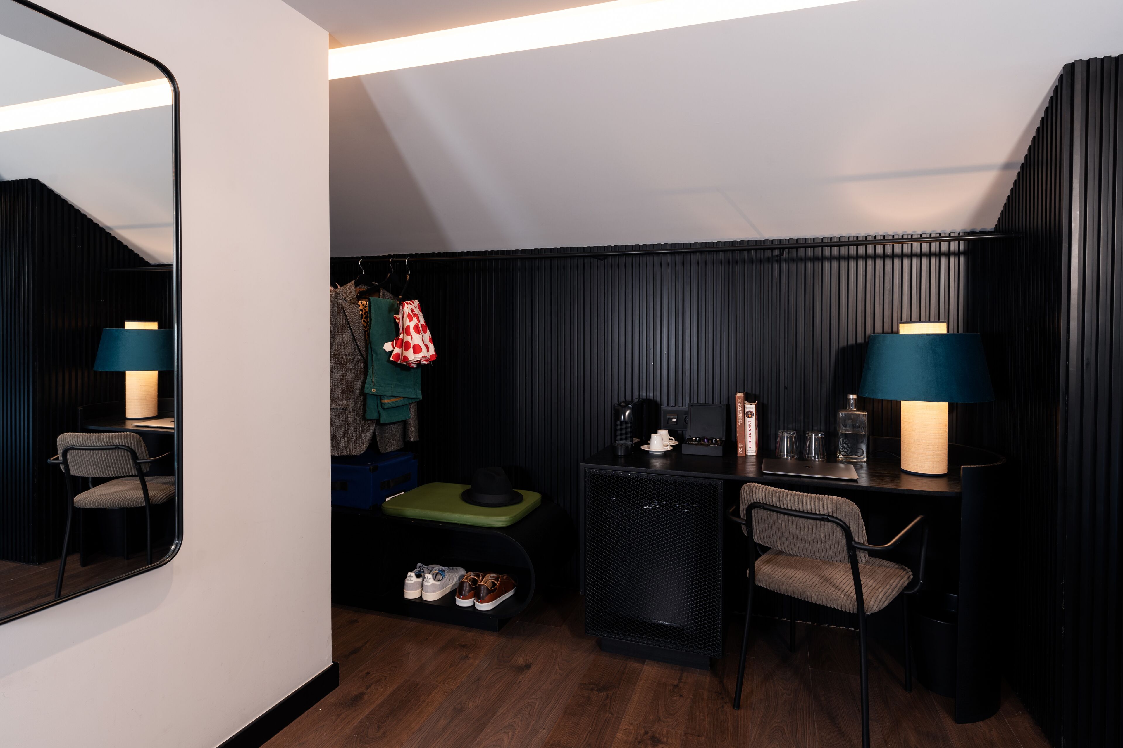 A compact hotel room with a mirrored wall, a small desk with a chair, a green ottoman, a coat rack with clothes, and a coffee machine.