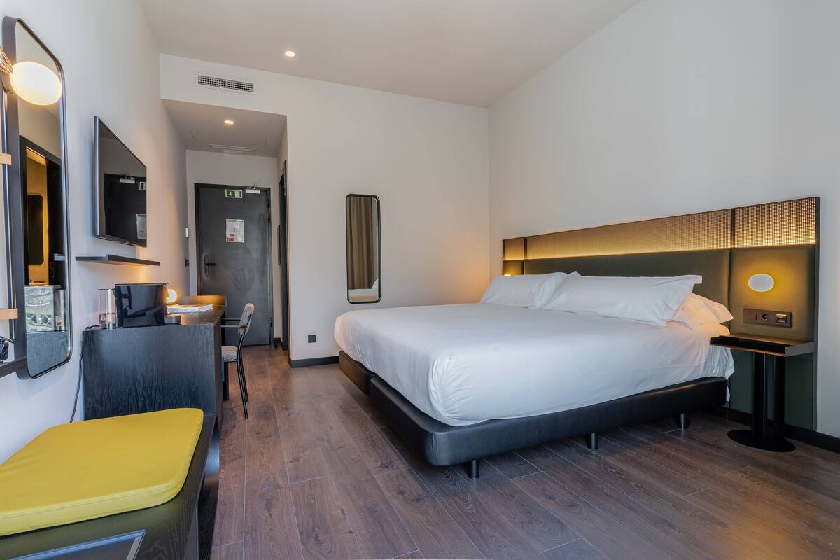 A modern hotel room with a large bed, a desk with a chair, a small seating area, and a mirror.
