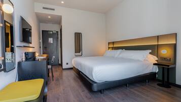Quarto de hotel moderno com uma cama grande, uma escrivaninha com cadeira, um pequeno espaço de estar e um espelho.