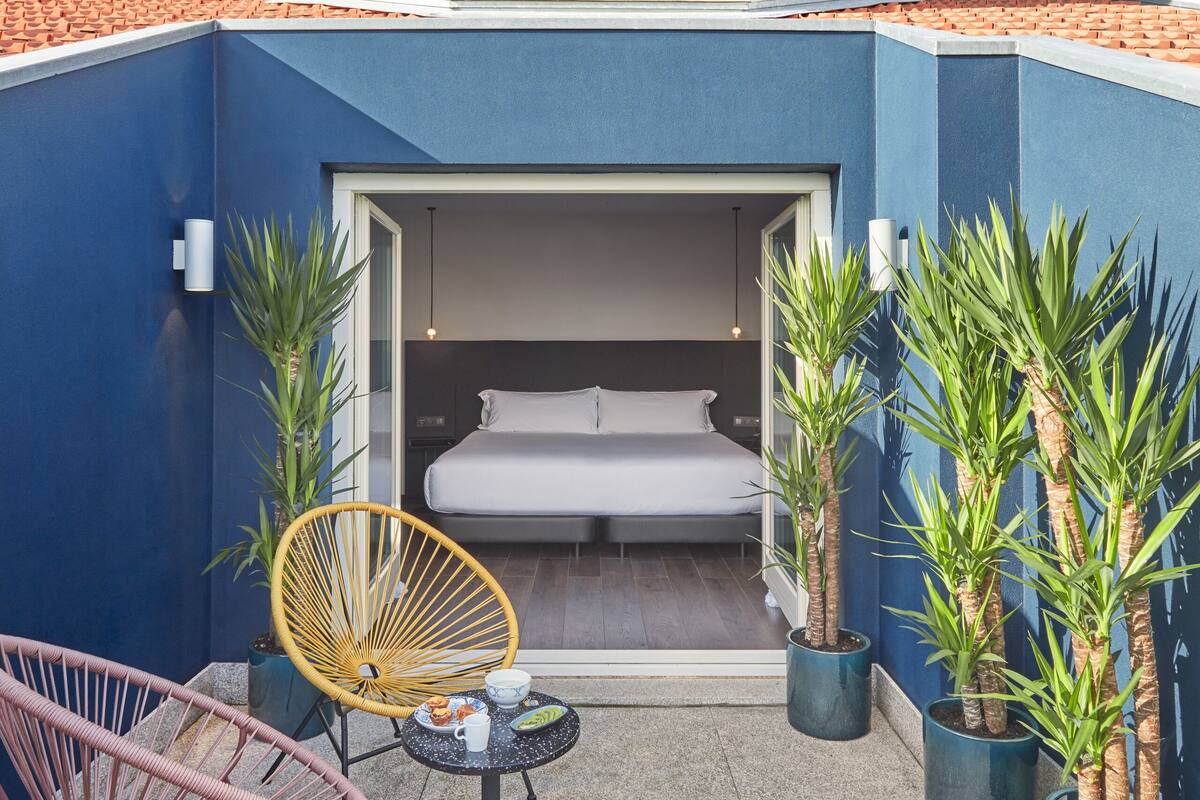 A compact, modern building with a white exterior and red-tiled roof. It features a small balcony with a bed, a yellow chair, and potted plants.