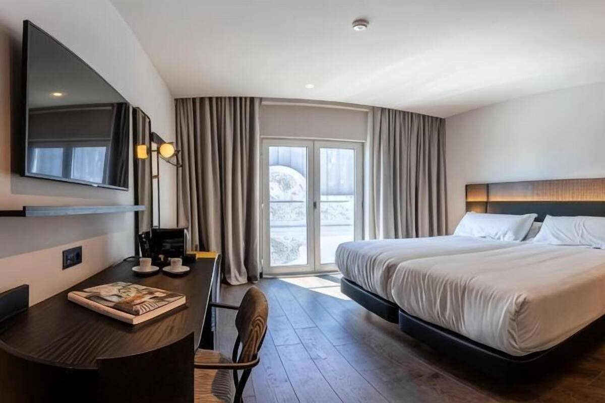 A modern hotel room with a bed, a desk with a chair, a flat-screen TV, and a large window with curtains.