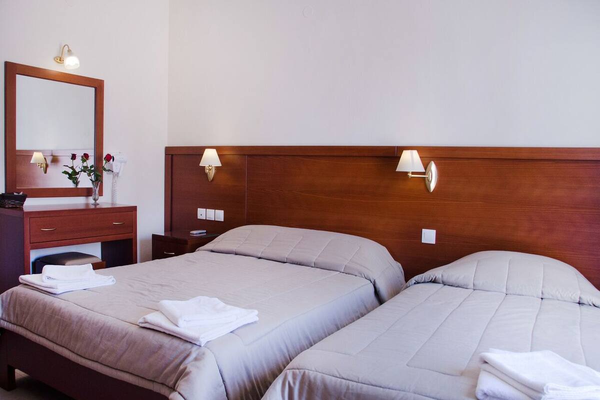 Habitación de hotel con dos camas, cabecera de madera, lámparas de noche, un espejo, tocador con teléfono y un jarrón con flores.