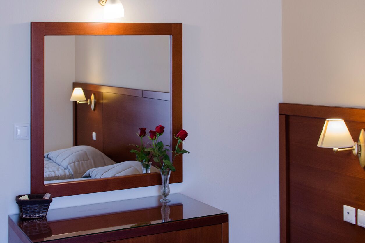 Habitación de hotel con cabecera de madera, un espejo grande, una mesita de noche con un jarrón de rosas rojas y una lámpara de pared.
